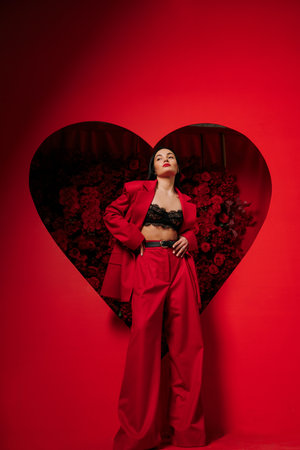 Beautiful girl in red pants and jacket posing on red background decorated by heart shape with red roses. Holiday party, birthday.の写真素材