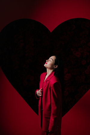Girl in red pants and jacket posing with closed eyes on red background decorated by heart shape. Holiday party, birthday. Side viewの写真素材