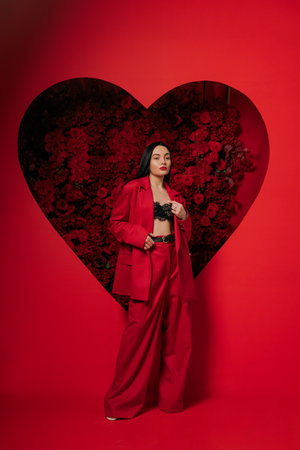 Beautiful girl in red pants and jacket posing on red background decorated by heart shape with red roses. Valentine's day. Holiday party, birthday.の写真素材