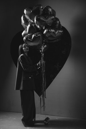 Beautiful girl in pants and jacket posing against big black heart with heart shape air balloons in her hands. Holiday party, birthday. Black and white image.の写真素材