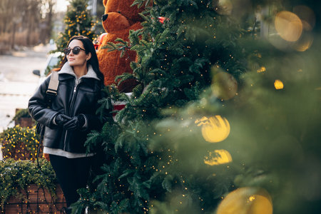 Young pretty woman in jacket and sunglasses standing on city street near Christmas tree decorated by glowing garlands. Image with bokeh effect.の写真素材