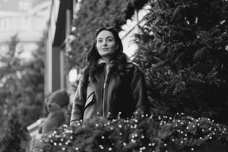 Young pretty woman in jacket standing on city street near Christmas tree decorated by glowing garlands. Black and white image.の写真素材