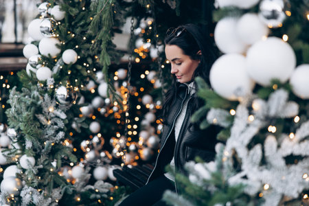 Young woman in jacket standing on city street near Christmas tree decorated by glowing garlands and balls.の写真素材