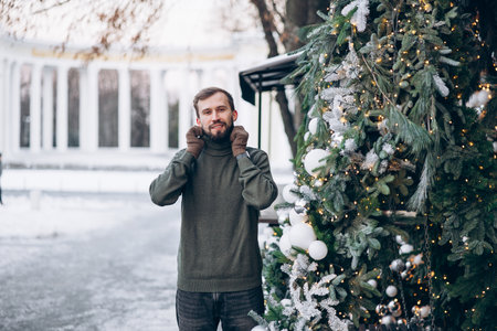 Young smiling bearded man in sweater and gloves standing on city street near Christmas tree decorated by balls and glowing garlands.の写真素材