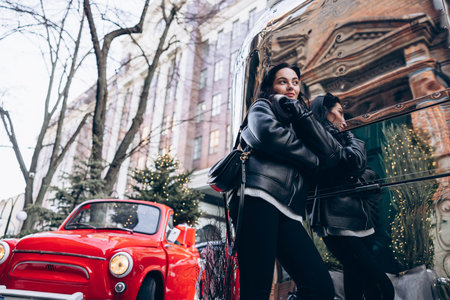 Young pretty woman in jacket with handbag standing on city street near herself reflection in big mirror against background of red retro car and Christmas tree decorated by glowing garlands.の写真素材