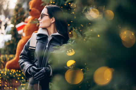 Portrait of young pretty woman in jacket and sunglasses standing on city street near Christmas tree decorated by glowing garlands. Image with bokeh effect.の写真素材