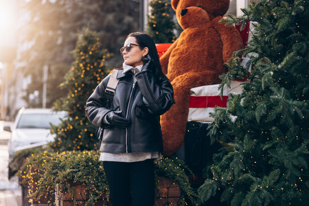 Young pretty woman in jacket and sunglasses standing on city street near Christmas tree decorated by glowing garlands.の写真素材