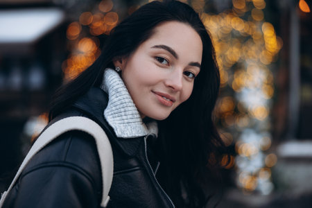 Portrait of young smiling woman on city street near Christmas tree decorated by glowing garlands. Image with bokeh effect.の写真素材