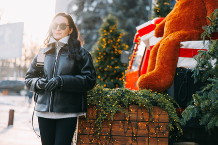 Young pretty woman in jacket and sunglasses standing on city street near Christmas tree decorated by glowing garlands. Backlit.の写真素材