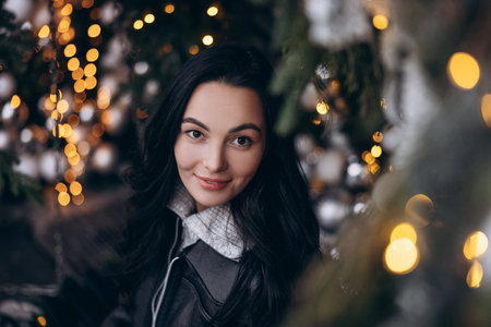 Portrait of young smiling woman on city street near Christmas tree decorated by glowing garlands. Image with bokeh effect.の写真素材