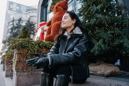 Young pretty woman in jacket sitting and resting on city street near Christmas tree decorated by glowing garlands.の写真素材