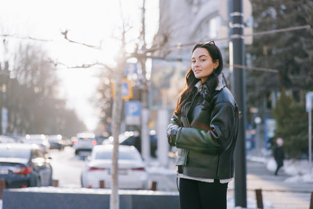 Young pretty brunette woman in jacket standing on city street against background of cars.の写真素材