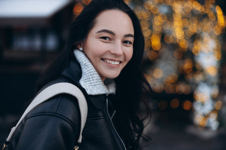 Portrait of young smiling woman on city street near Christmas tree decorated by glowing garlands. Image with bokeh effect.の写真素材