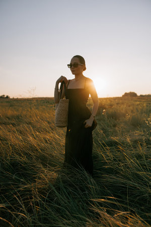 Young pretty woman in black dress and sunglasses walking on meadow through herbs at sunset. Backlight.の写真素材