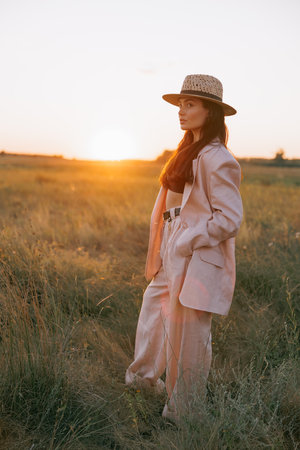 Young pretty woman in jacket, pants and straw hat walking on meadow through herbs at sunset. Backlight.の写真素材