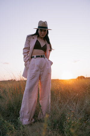 Young pretty woman in black jacket, pants and straw hat walking on meadow through herbs at sunset. Backlight.の写真素材