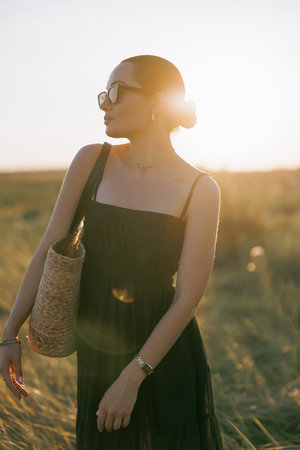 Young pretty woman in black dress and sunglasses walking on meadow through herbs at sunset. Close-up. Backlight.の写真素材