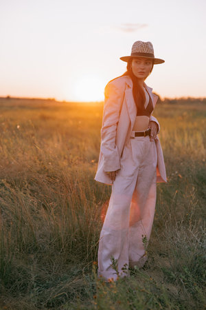 Young pretty woman in jacket, pants and straw hat walking on meadow through herbs at sunset. Backlight.の写真素材