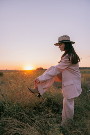 Young pretty woman in jacket, pants and straw hat walking on meadow through herbs at sunset. Backlight.の写真素材