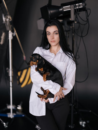 Young brunette woman in white shirt posing in studio for shooting holding her dachshund dog on her hands against background of spotlights and tripods. Close-up.の写真素材