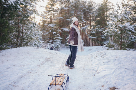Happy young woman pulling the sled uphill in forest during walk among snow covered pine trees in...