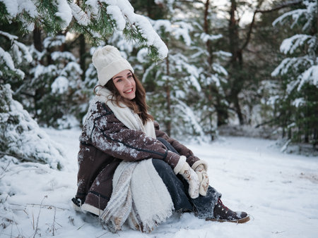 Happy young smiling woman sitting on ground and enjoying during forest walk among snow covered pine trees in winter day.の写真素材