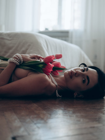 Brunette woman lying on the floor near bed with bouquet of red tulips in pearl necklace and bracelet. Close-up.の写真素材