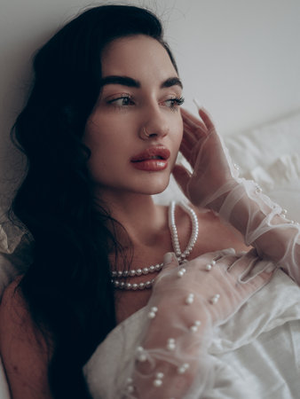 Portrait of beautiful pensive brunette woman lying in bed in bridal transparent gloves and pearl necklace. Wedding day, preparations. Close-up.の写真素材
