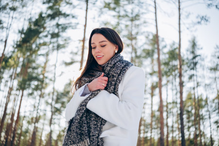 Portrait of young brunette woman in white coat and scarf among pine trees during walk in winter day.の写真素材