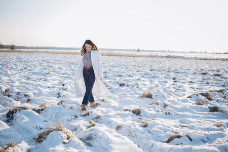 Happy young brunette woman walking on snow covered meadow among dry grass tussocks in winter sunny day.の写真素材