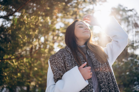 Portrait of young brunette woman in white coat and scarf enjoying with closed eyes by pine forest in winter day. Backlight.の写真素材