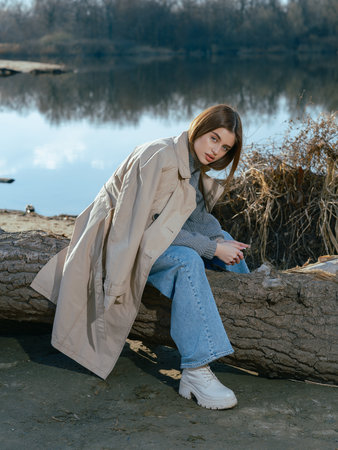 Young woman in trench coat sits on log on beach by lake with calm waters reflecting the sky and clouds in early spring. Close-up.の写真素材