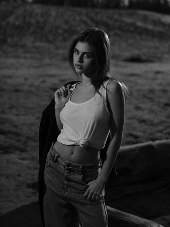Young brunette woman in white tank top and jeans holding jacket in her hands and standing near logs on sand beach. Close-up. Black and white image.の写真素材