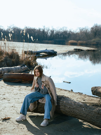 Young woman in trench coat sits on log on beach by lake with calm waters reflecting the sky, clouds and surrounded by bare trees in early spring.の写真素材