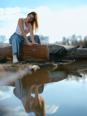 Young woman in white tank top and jeans sitting and relaxing on log on beach by lake with calm waters reflecting sky with clouds and herself.の写真素材