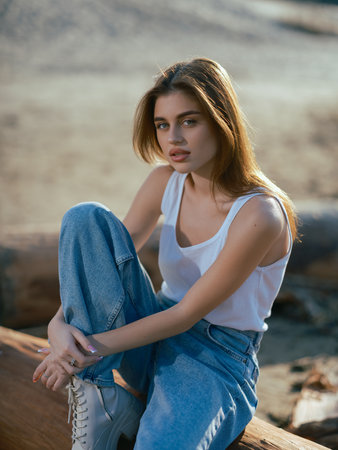 Portrait of young brunette woman in white tank top and jeans sitting on log on sand beach.の写真素材