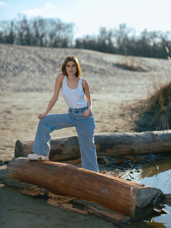 Young brunette woman in white tank top and jeans standing near log on beach against background of sand hill and sky.の写真素材
