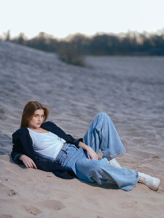 Young brunette woman in white tank top, jeans and jacket lies down on beach against background of sand and sky.の写真素材