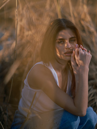 Portrait of young brunette woman in white tank top and jeans sitting and relaxing among dry reed. Close-up.の写真素材