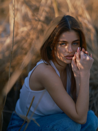 Portrait of young brunette woman in white tank top and jeans sitting and relaxing among dry reed. Close-up.の写真素材