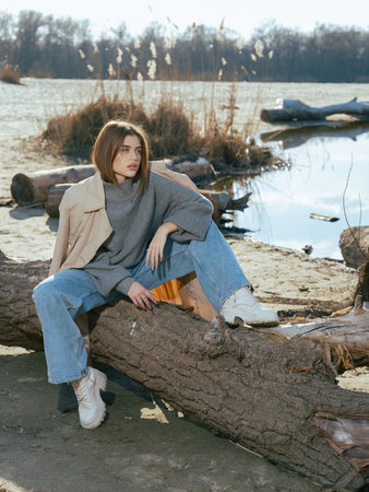 Young woman in trench coat sits on log on beach by lake with calm waters reflecting the sky and clouds in early spring.の写真素材