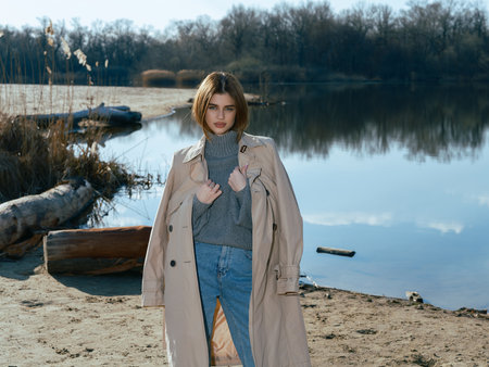 Young woman in trench coat stands on beach by lake with calm waters reflecting the sky, clouds and surrounded by bare trees in early spring.の写真素材