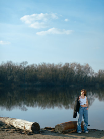 Young woman in white tank top and jeans holding jacket and stands on beach by logs and lake with calm waters reflecting the sky, clouds and surrounded by bare trees in early spring.の写真素材