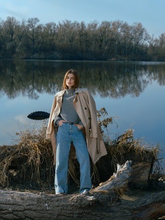 Young woman in trench coat stands on beach by logs and lake with calm waters reflecting the sky, clouds and surrounded by bare trees in early spring.の写真素材