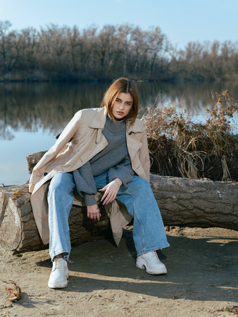 Young woman in trench coat sits on log on beach by lake with calm waters reflecting the sky, clouds and surrounded by bare trees in early spring.の写真素材