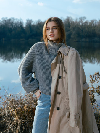 Young woman with trench coat stands by lake with calm waters reflecting the sky, clouds and surrounded by bare trees in early spring. Close-up.の写真素材