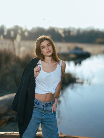 Young woman in white tank top and jeans holding jacket and stands on beach by lake with calm waters reflecting the sky and clouds. Close-up.の写真素材