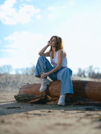 Young brunette woman in white tank top and jeans sits on log on sand beach against background sky with clouds.の写真素材