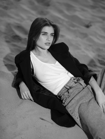 Young brunette woman in white tank top, jeans and jacket lies down on beach against background of sand. Top view. Black and white image.の写真素材