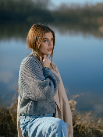 Young pensive woman in sweater stands by lake with calm waters reflecting the sky and clouds in early spring. Close-up.の写真素材
